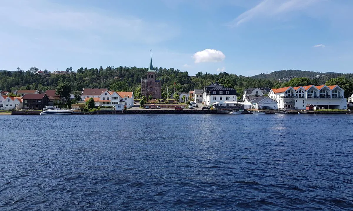 Kommunal gjestebrygge Svelvik sentrum: Gjestebryggen strekker seg fra den åpne plassen med parkerte biler og mot høyre billedkant.
Restaurant Dreggen omtrent midt i bildet og Kiwi i stort bygg med rød takstein.
Ankerdrammen kjøpes på senteret etter man har passert fergeleiet ved Lallaparken.